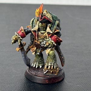 Pode incluir: Uma figura em miniatura de uma criatura com tema de pirata, com um corpo reptiliano verde e amarelo, um chapéu de três pontas e um casaco vermelho e dourado. A figura segura uma espada e está sobre uma base marrom, mostrando detalhes de pintura e um tema de fantasia.
