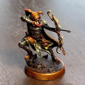 May include: A painted miniature figurine of an elf archer, wearing a gold and silver armor, holding a bow and arrow. The figurine is standing on a brown base with a gold rim.
