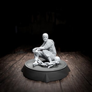 May include: A grayscale sculpture of a seated man with a beard, wearing textured clothing, and resting on a round platform. A skull and chain are near his feet. The sculpture is set against a dark background.