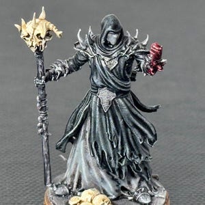 Könnte beinhalten: Miniaturfigur eines verhüllten Zauberers mit einem Stab mit Totenkopf und einer roten, klauenartigen Hand. Die Figur ist in Schwarz-, Grau- und Goldtönen bemalt und steht auf einem detaillierten Sockel. Ein Fantasy-Sammlerstück.