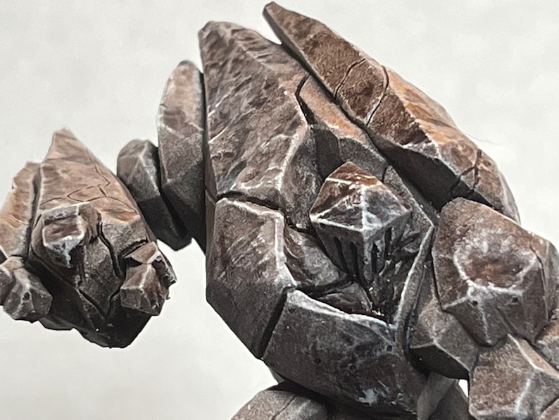 Pode incluir: Um close-up de uma figura de golem de pedra cinza, marrom e branca. O golem &eacute; feito de muitas pe&ccedil;as de pedra diferentes e tem uma superf&iacute;cie &aacute;spera e texturizada.