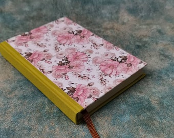 May include: A small, floral-patterned notebook with a yellow spine and a brown ribbon bookmark. The cover features a watercolour-style design of pink roses and other flowers on a white background.