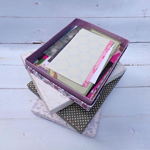 Puede incluir: Una pila de cajas decorativas de varios tamaños y patrones. La caja superior, abierta, revela una colección de tarjetas y papeles estampados. Las cajas presentan diseños florales, de lunares y abstractos en tonos blancos, verdes y morados.