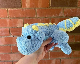May include: A light blue crochet dragon with yellow accents. The plush toy has black eyes and a textured, fluffy appearance. The dragon is held against a brick wall background.
