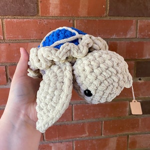 May include: A crocheted stuffed turtle toy with a blue shell and beige body. The turtle has black eyes and a small white tag attached to its shell.