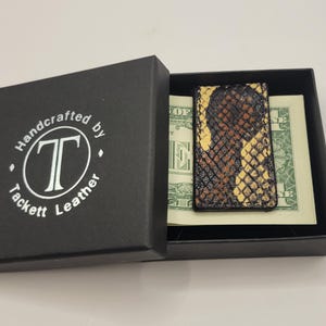 May include: A rectangular money clip with a textured, snakeskin-patterned finish in shades of black, brown, and yellow. The clip holds a one-dollar bill inside a black gift box featuring the white text Handcrafted by Tackett Leather.