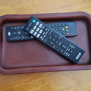 May include: Two black rectangular remote controls with numerous buttons and text labels resting inside a brown, smooth, rectangular tray with rounded corners and raised edges. The top remote features a green power button and white text.