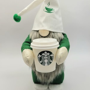 May include: A green and white gnome holding a Starbucks coffee cup. The gnome is wearing a white hat with a green coffee cup design and a green pom-pom.
