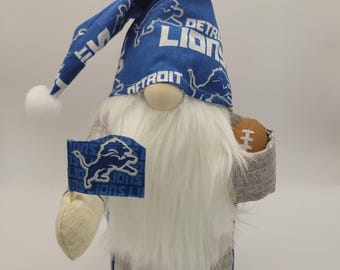 May include: A plush gnome wearing a blue and white Detroit Lions hat, a grey shirt, and holding a brown football. The gnome has a white beard and blue and white feet.
