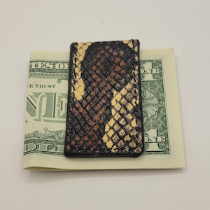 May include: A rectangular money clip featuring a textured, snakeskin-patterned surface in shades of black, brown, and cream. The clip is constructed with dark stitching along the edges and is shown holding a single United States one-dollar bill.