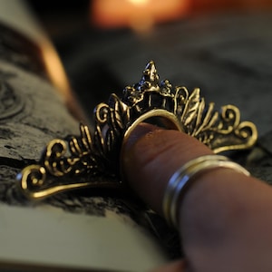 Op de afbeelding: Een goudkleurige ring met een sierlijk kroonontwerp, gedragen aan een vinger. De ring heeft ingewikkelde blad- en werveldetails. Extra zilveren ringen zijn ook zichtbaar. De achtergrond bevat een wazig boek en kaarsen.