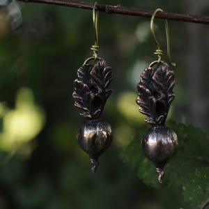 Może przedstawiać: Para ręcznie robionych kolczyków o tematyce botanicznej. Każdy kolczyk ma ciemny, brązowy wzór liścia nad zaokrągloną, metaliczną perłą. Kolczyki wiszą na złotych drucikach, na rozmytym zielonym tle.