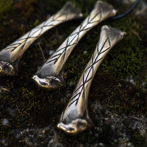 Könnte beinhalten: Drei bronzefarbene Anhänger in Knochenform mit geätzten geometrischen Mustern. Jeder Anhänger hängt an einer schwarzen Kordel und liegt auf einer moosigen Oberfläche. Die Anhänger haben ein verwittertes, antikes Aussehen.