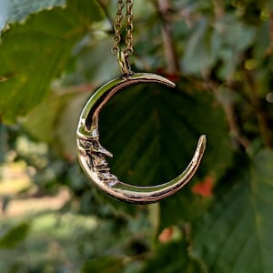 Peut inclure: Un pendentif en forme de croissant de lune en métal doré suspendu à une fine chaîne à maillons assortie. La lune présente un profil de visage humain en relief détaillé sur sa surface, avec une finition polie et réfléchissante.