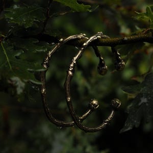 Pode incluir: Duas pulseiras douradas em forma de galho são exibidas em um galho de árvore com folhas verdes. As pulseiras têm um design texturizado e orgânico com pequenos enfeites redondos. O fundo é uma visão desfocada de mais folhagem verde.