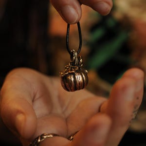 Peut inclure: Un pendentif en forme de citrouille en bronze, suspendu à un cordon noir. Le pendentif présente des lignes verticales et une tige texturée. Le bijou est tenu dans une main, soulignant le design détaillé et le thème automnal.