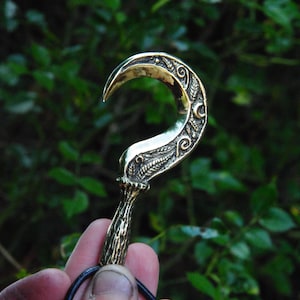 Op de afbeelding: Een bronzen haakvormige hanger met gedetailleerde bloemen- en bladerpatronen. Het gebogen blad is bevestigd aan een zwart koord. De achtergrond is een vage weergave van groen gebladerte, wat een buitenomgeving suggereert.