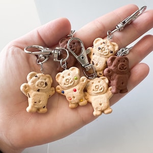 Könnte beinhalten: Ein Set mit sechs Schlüsselanhängern in Form von Keksbären. Die Bären sind aus Polymer Clay gefertigt und bemalt, um wie verschiedene Arten von Keksen auszusehen. Es gibt zwei Schokoladenkeksbären, einen Schokoladenkeksbär, einen Streuselkeksbär und zwei einfache Keksbären.