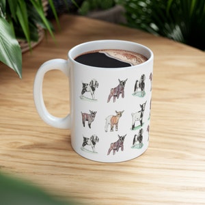 May include: White ceramic coffee mug with a black handle. The mug is decorated with a pattern of black and white goats. The goats are drawn in a cartoon style and are all different sizes and poses.