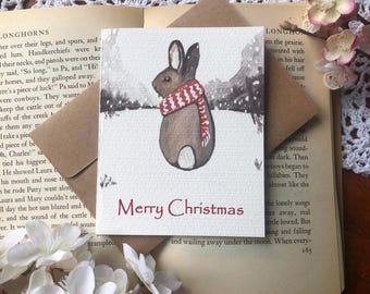 May include: A watercolor Christmas card featuring a brown rabbit wearing a red and white scarf, set against a snowy backdrop. The words "Merry Christmas" are written in red. Includes a brown envelope.