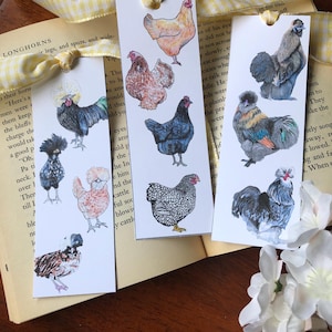 May include: Three rectangular bookmarks featuring watercolor illustrations of chickens in various breeds and colors. The bookmarks are arranged on an open book with a yellow ribbon. The illustrations include a variety of chicken breeds.