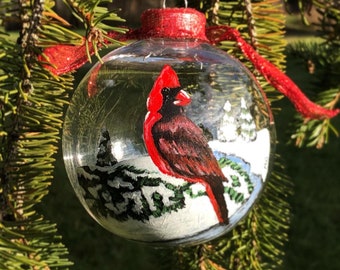 May include: A clear glass Christmas ornament with a hand-painted cardinal bird scene. The cardinal is red and black, perched on a snowy branch. The ornament has a red glitter top and is hanging from a white ribbon.