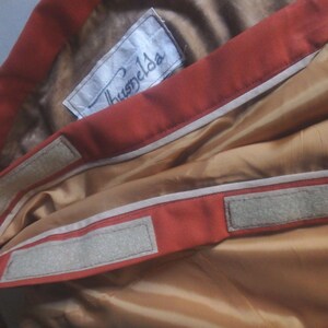 Op de afbeelding: Close-up van de binnenkant van een rood jack met een wit label met de tekst "Huspels" en twee witte klittenbandstrips.