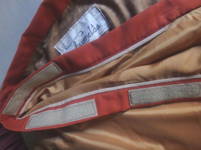 Op de afbeelding: Close-up van de binnenkant van een rood jack met een wit label met de tekst "Huspels" en twee witte klittenbandstrips.