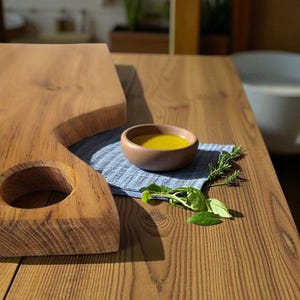 Può includere: Un tagliere in legno con un ritaglio circolare e una piccola ciotola di legno piena di liquido giallo. La ciotola è appoggiata su un panno blu, con rametti di rosmarino e basilico nelle vicinanze. Il legno ha una venatura naturale.