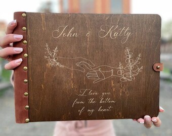 May include: A brown wooden photo album with a leather cover and brass studs. The album is personalized with the names "John & Katty" and the inscription "I love you from the bottom of my heart."