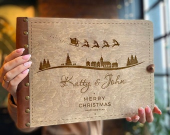 Könnte beinhalten: Ein Holzfotoalbum mit braunem Lederbezug und Metallnieten. Das Album zeigt ein lasergraviertes Design von Santa Claus, der über ein verschneites Dorf fliegt, mit dem Text "Katy & John Frohe Weihnachten Frohes Neues Jahr".