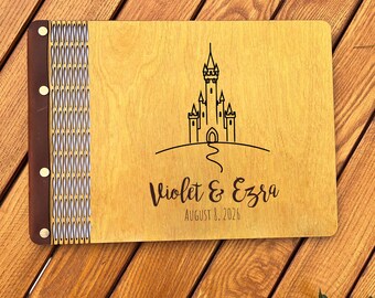 May include: A wooden wedding guest book with a castle illustration and the names "Violet & Ezra" and the date "August 3, 2026" engraved on the cover. The book is next to a stack of wedding photos and a pen.
