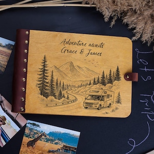 Könnte beinhalten: Ein Fotoalbum aus Holz mit einem braunen Leder-Rücken und Verschluss. Der Einband ist mit einer Berg- und Wohnmobil-Szene graviert, darüber steht "Adventure awaits Grace & James". Fotos einer Safari sind um das Album verstreut.