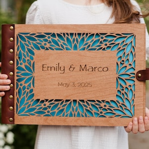 May include: A wooden photo album with a teal blue floral design and the text "Emily & Marco" and "May 3, 2025" engraved on the front.