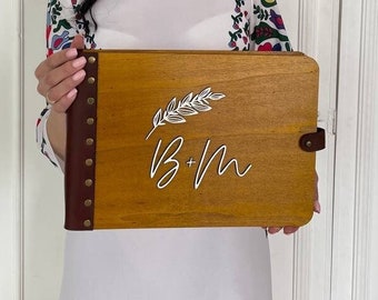 Op de afbeelding: Een vrouw houdt een houten fotoalbum vast met een bruine leren kaft en een wit verenontwerp. Het album heeft de initialen "B+M" gegraveerd op de voorkant.