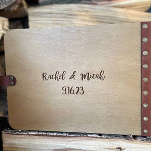 K&ouml;nnte beinhalten: Ein G&auml;stebuch aus Holz mit einem braunen Lederr&uuml;cken und Messingnieten. Der Einband ist mit dem Text "Rachel & Micah 9.16.23" graviert.