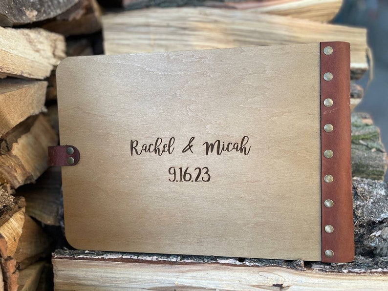 K&ouml;nnte beinhalten: Ein G&auml;stebuch aus Holz mit einem braunen Lederr&uuml;cken und Messingnieten. Der Einband ist mit dem Text "Rachel & Micah 9.16.23" graviert.