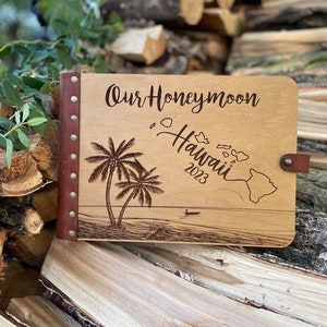 K&ouml;nnte beinhalten: Ein Holzfotoalbum mit braunem Lederbezug und Metallnieten. Das Album ist mit dem Text "Our Honeymoon Hawaii 2023" graviert und zeigt ein Palmen- und Inselmotiv.