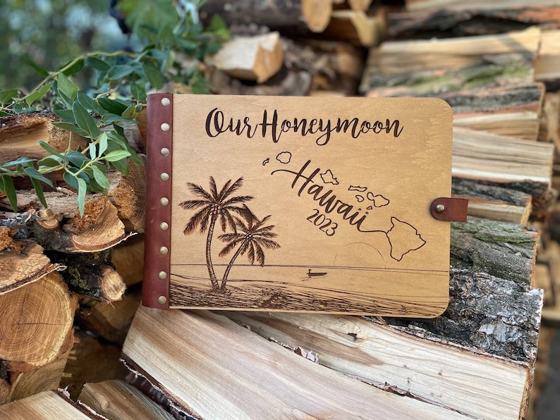 K&ouml;nnte beinhalten: Ein Holzfotoalbum mit braunem Lederbezug und Metallnieten. Das Album ist mit dem Text "Our Honeymoon Hawaii 2023" graviert und zeigt ein Palmen- und Inselmotiv.