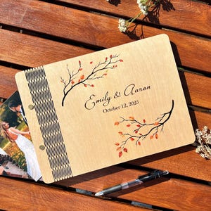 May include: A wooden photo album with the names "Emily & Aaron" and the date "October 12, 2025" engraved on the cover. The cover features a branch design with orange leaves. A photo is visible inside the album.