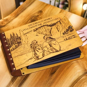 May include: A brown leather photo album with a wooden cover engraved with the text "Iacobucci Family Yellowstone Adventures" and a scene of two people hiking in a forest. The album has metal rivets along the spine.