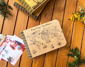 May include: A wooden travel memories journal with a world map design and the text "Murray & Lyn's Travel Memories." The journal is open, with photos and other journals nearby. The background is a wooden surface.