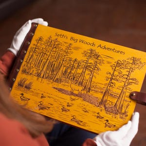 Puede incluir: Un diario encuadernado en cuero amarillo con el texto "Seth's Big Woods Adventures" grabado en la portada. La portada presenta una escena de naturaleza con árboles, un ciervo y patos.