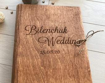 Op de afbeelding: Een houten trouwalbum met de woorden "Bilenchuk Wedding" en de datum "15.08.20" gegraveerd op de omslag. Het album is vastgebonden met een touwstrik. Het hout heeft een warme, roodbruine tint.