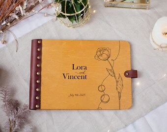 May include: A wooden guest book with a floral design and the names "Lora and Vincent" and the date "July 4th 2025" printed on the cover. The book has a brown leather spine with gold rivets and a leather strap closure.