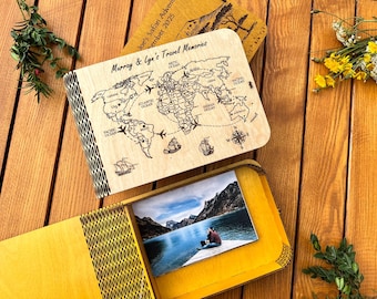 Może przedstawiać: Drewniane pudełko na pamiątki z podróży z mapą świata i napisem "Murray & Lyn's Travel Memories". Pudełko jest otwarte, ukazując zdjęcie osoby nad jeziorem. Tło obejmuje drewnianą powierzchnię i elementy dekoracyjne.