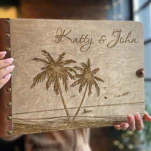 Può includere: Un album fotografico in legno con una copertina in pelle marrone e un design inciso a laser di palme e il testo "Katty & John".