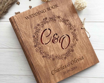 Op de afbeelding: Een houten trouwboek met een bloemenkransontwerp en de initialen "C&O" in het midden. De namen "Charles & Olivia" en de datum "21.09.2019" zijn gegraveerd onder de krans.