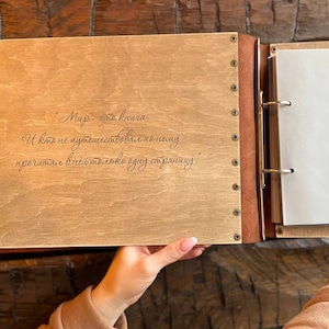 K&ouml;nnte beinhalten: Ein Holzfotoalbum mit braunem Lederr&uuml;cken und Metallringen. Der vordere Einband hat ein in Russisch eingraviertes Zitat: "Die Welt ist ein Buch. Und wer nicht durch sie gereist ist, hat nur eine Seite gelesen."