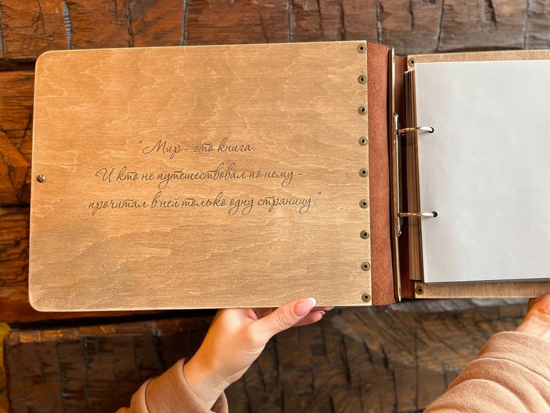 K&ouml;nnte beinhalten: Ein Holzfotoalbum mit braunem Lederr&uuml;cken und Metallringen. Der vordere Einband hat ein in Russisch eingraviertes Zitat: "Die Welt ist ein Buch. Und wer nicht durch sie gereist ist, hat nur eine Seite gelesen."